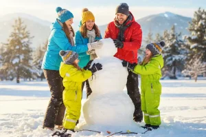 A family, warmly dressed in colorful winter gear, laughing together while building a snowman on a sunny, snowy day, capturing the fun and challenges of outdoor winter activities with children.