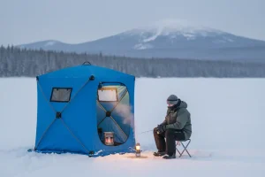 A portable, insulated ice fishing shelter or shanty set up on a frozen, snow-covered lake, with an angler tending to a fishing rod inside, highlighting the comfort and dedicated focus of the winter sport.