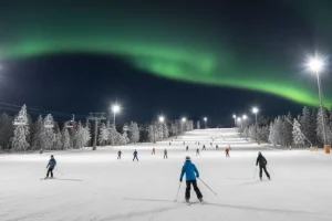 The main ski slope at Levi, Finland, illuminated by floodlights under a dark Arctic sky, with the faint green glow of the Aurora Borealis visible.
