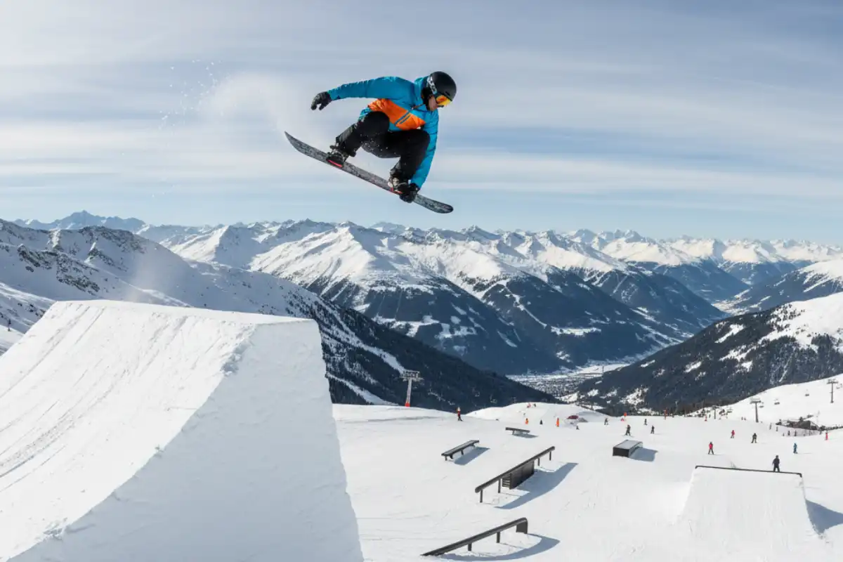 A snowboarder launching off a jump in a snow park on the Jakobshorn mountain in Davos, with the Alps visible in the background.