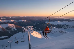 A solo skier riding a chairlift up a snowy mountain slope with vast panoramic views