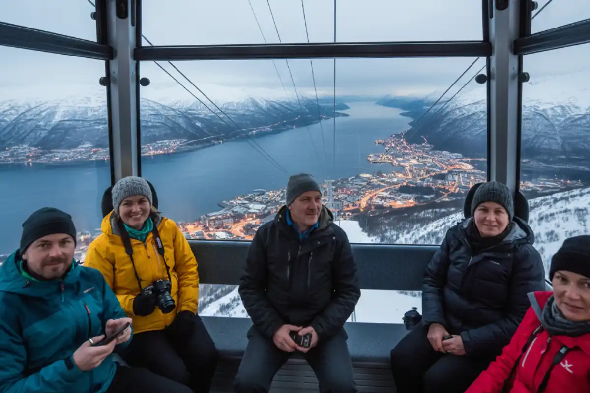 Tourists riding the Narvik Fjellheisen gondola up the mountain, with a breathtaking panoramic view of the deep blue fjord, the city, and snow-covered peaks.