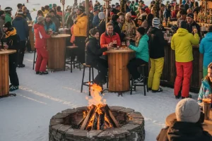 People celebrating at a lively outdoor bar at the base of a snowy mountain during the late afternoon
