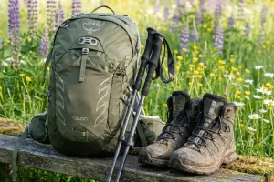 A well-organized spread of modern hiking equipment including a lightweight backpack, trekking poles, and waterproof boots set against a background of spring wildflowers