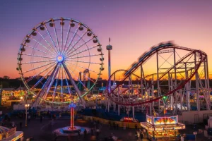 A vibrant amusement park skyline at dusk with a lit-up Ferris wheel and a high-speed roller coaster track against a purple sky