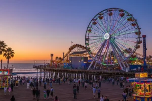 The solar-powered Ferris wheel and roller coaster of Pacific Park glowing against the twilight sky at the end of the wooden Santa Monica Pier