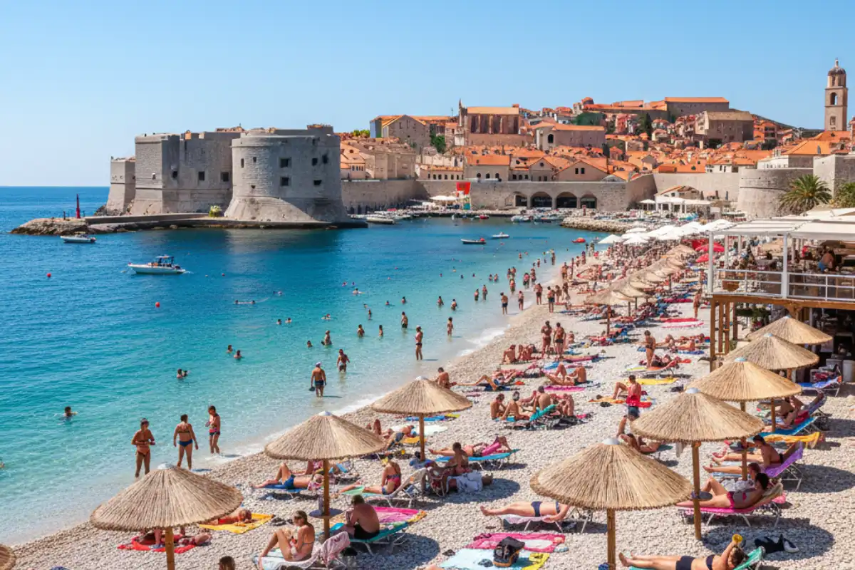 People relaxing on the pebbled Banje Beach with the historic stone fortifications of Dubrovnik rising in the immediate background