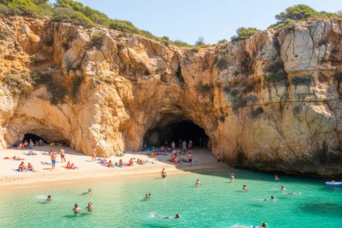 Two small sandy beaches connected by a tunnel through a jagged cliff face with people swimming in vibrant blue water