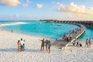 A string of luxury timber overwater villas extending into a vibrant turquoise lagoon with a white sand sandbar visible in the distance.